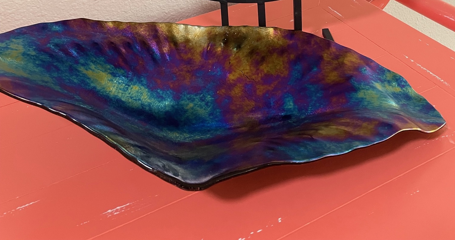 Tide Platter: an iridescent wavy slumped fused-glass platter in shifting blues, purples, teals, and gold, resting on a coral-colored table.