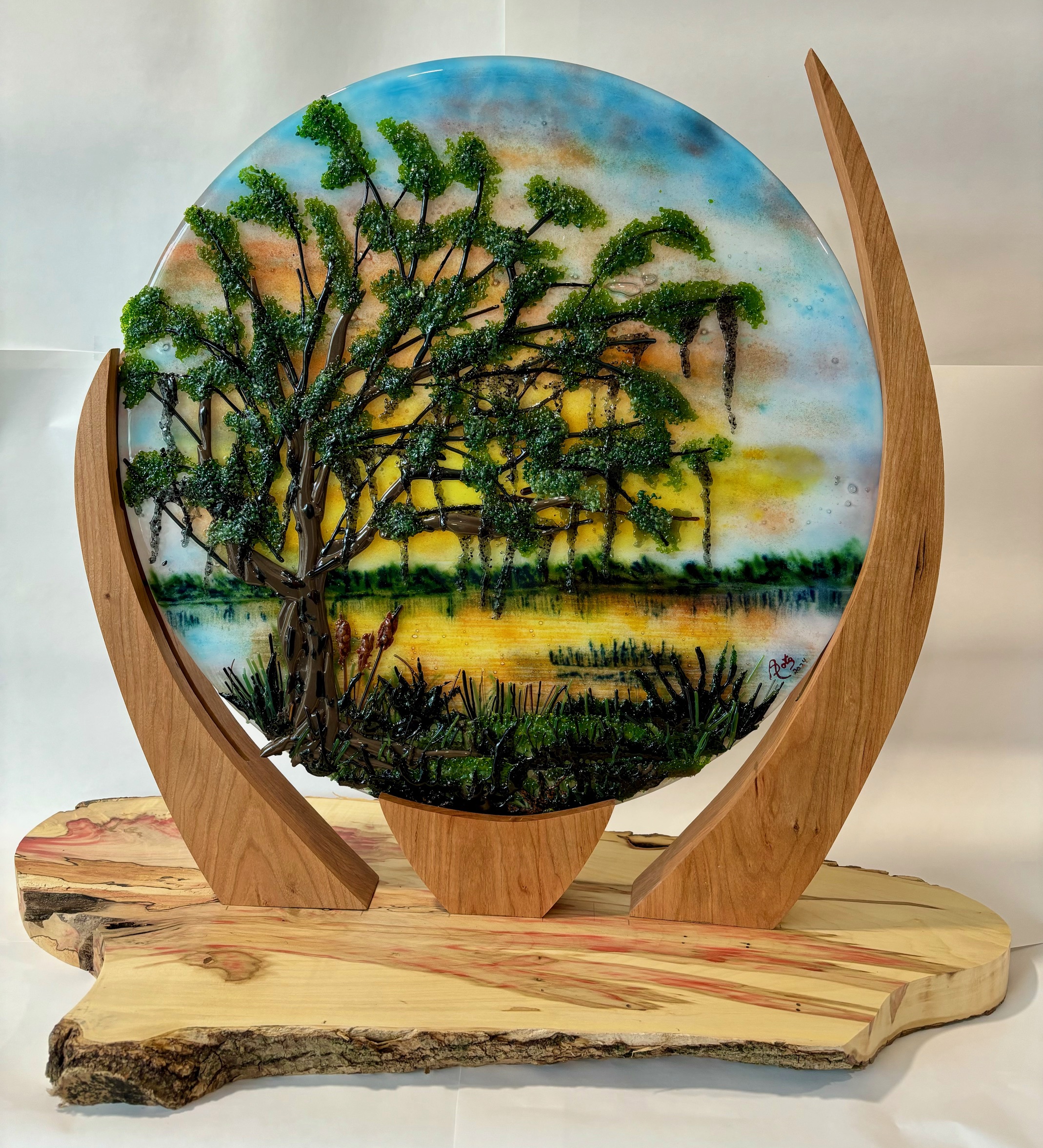 Circular fused-glass scene of an oak tree by a lake.